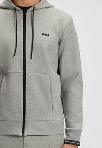 Felpa grigia realizzata in morbido tessuto con zip frontale, tasche anteriori, accenti neri e un piccolo logo sul petto. Design con colletto alto.