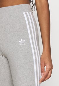 Grå leggings i mjukt tyg med tre vita ränder längs sidan och en vit Adidas-logga på höften.
