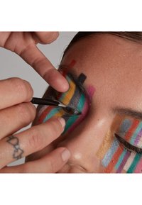 Ett livfullt ansiktsmakeup med färgglada ränder i blått, gult, rosa och svart. Närbild på ögat med en hand som håller en borste.