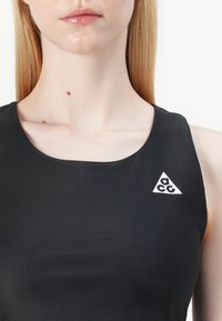 Melna bezroku blūze ar apaļu kakla izgriezumu un baltu trīsstūra logotipu kreisajā pusē, izgatavota no gludas, elastīgas auduma.