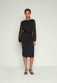Čierne šaty s dlhými čipkovými rukávmi, úzky strih, dĺžka po kolená a detail s viazaním vpredu v oblasti pása. Hladká látka s textúrovanými akcentmi.