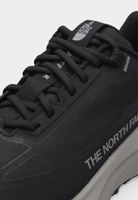 Μαύρο αθλητικό παπούτσι The North Face με χοντρές μαύρες κορδόνια, υφασμάτινο επάνω μέρος με υφή και λευκή σόλα, με το εμπορικό λογότυπο στο πλάι και στη γλώσσα.