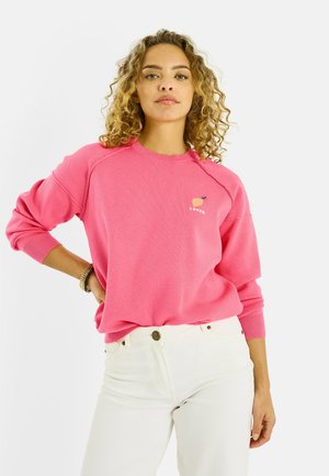 Mujer joven con cabello rizado, vistiendo una sudadera rosa con un pequeño gráfico de durazno y pantalones blancos, posando con una mano en la cadera.