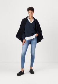 Marineblå oversized genser lagt over en stripete skjorte med krage, kombinert med lyseblå skinny jeans og svarte loafers.