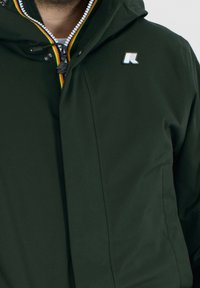 Ciemnozielona kurtka z zamkiem błyskawicznym z przodu, kapturem i wielokolorową lamówką przy zamku. Na piersi znajduje się małe niebieskie logo. Gładka faktura tkaniny.