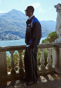 Fekete sportos dzseki kék részletekkel, fekete nadrággal, amely fehér csíkokkal van díszítve. A modell egy kőkorlátnál áll, ahonnan a vízre néz.