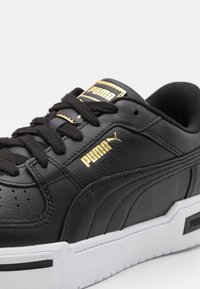 Zapatilla de cuero negro de Puma con logo dorado, cordones negros y suela blanca, mostrada en primer plano sobre un fondo blanco.