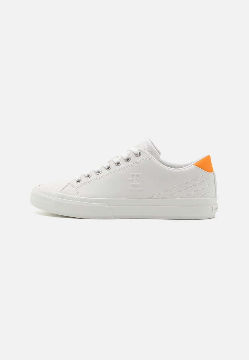 Basket blanche basse avec lacets blancs, logo subtilement embossé sur le côté et languette de talon orange sur un fond clair uni.
