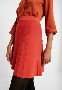 Jupe plissée rouge avec une ceinture ajustée, confectionnée dans un tissu texturé. Elle présente un petit accent de marque sur la ceinture pour plus de détails.