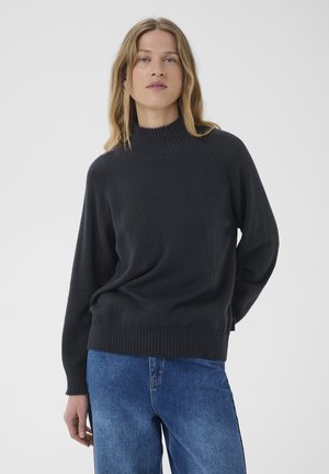 Femme aux cheveux châtain clair portant un pull noir à col montant et un jean bleu, debout devant un fond clair uni.