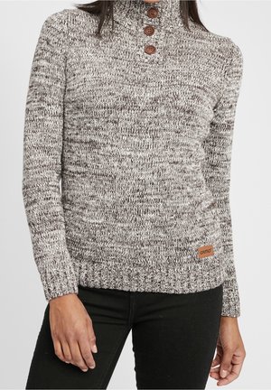 Femme portant un pull en tricot gris et blanc avec un col montant et trois boutons en bois, associé à un pantalon noir.