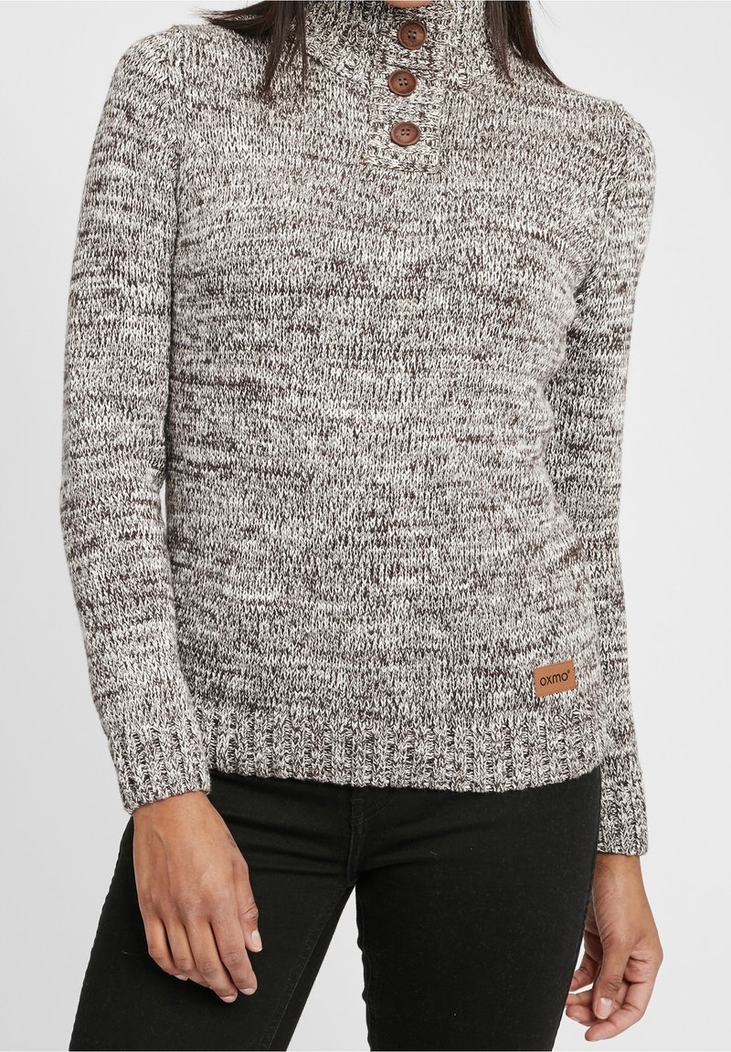 Femme portant un pull en tricot gris et blanc avec un col montant et trois boutons en bois, associé à un pantalon noir.