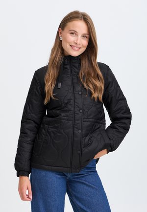 Femme aux longs cheveux châtain clair, portant une veste matelassée noire et un jean bleu, souriant avec une main dans la poche.