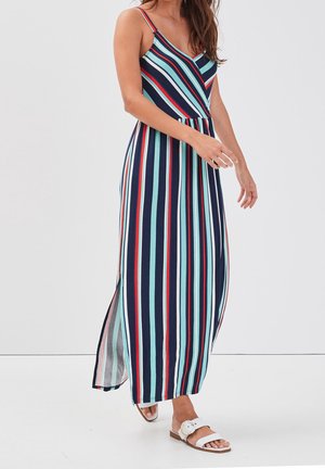 Vrouw die een mouwloos maxijurk draagt met verticale strepen in marineblauw, lichtblauw, rood en wit, lopend op witte sandalen.
