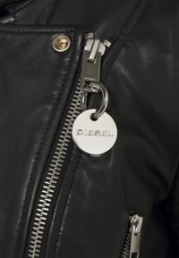 Černá kožená bunda s stříbrnými detaily, obsahující výrazný zip, klopu na knoflíky a kulatou visačku s vyrytým nápisem "DIESEL."