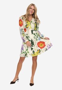 Smilende kvinde i en blomstret, langærmet kjole, der holder i skørtet, iført sorte, hælede slingback-sko mod en ensfarvet baggrund.