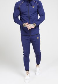 Navy tracksuit comprising a zip-up jacket and fitted pants, accented with gold stripes and logo. Features ribbed cuffs and smooth fabric.