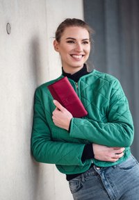 Green quilted jacket with a zip, black turtleneck, and a red wallet. Wall in the background, person leaning against it, smiling.