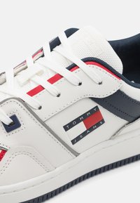 Zapatilla blanca de cuero con acentos en rojo y azul marino, que presenta un upper texturizado, lengüeta acolchada y un logo de "TOMMY JEANS" en el lateral.