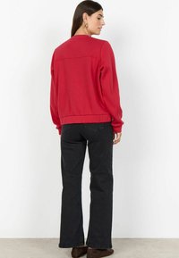 Felpa rossa con scollo rotondo e orlo a coste, abbinata a jeans neri a zampa. La modella mostra la vista posteriore, capelli sciolti e accessori minimal.