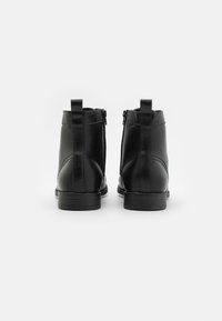 Botas de cuero negro hasta el tobillo con una textura suave, cremalleras laterales y tiradores en la parte posterior. Las suelas son planas y ligeramente texturizadas.
