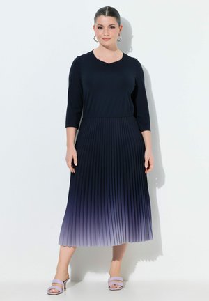 Kvinde med mørkt hår i en glat knold iført en marineblå, trekvartærmet plisseret kjole, der toner ud i lyse lilla ved kanten, og lyse lilla sandaler.