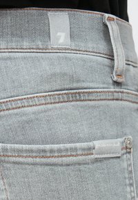 Blugi din denim gri cu o textură subtilă, cusături portocalii și o etichetă cu numărul 7. Include un detaliu mic de buzunar.