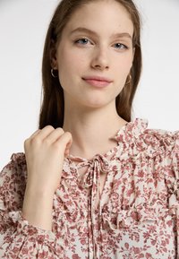Květinová blůza s volánovými akcenty ve světlé smetanové a tlumené růžové barvě. Disponuje zavazovacím výstřihem a lehkým materiálem s jemným vzorem.