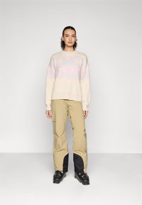 Helles beiges Sweatshirt mit pastellfarbenen geometrischen Mustern, kombiniert mit beigen Skihosen mit schwarzen Akzenten. Modell trägt schwarze Skischuhe.