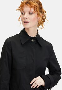 Schwarzer Trenchcoat mit spitzem Kragen, vorderem Knopfverschluss und seitlichen Taschen. Glattes Material mit minimalen Nähten und tailliertem Schnitt.