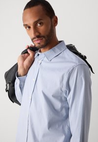 Man in een lichtblauwe geruite blouse met knopen, met een zwarte leren tas over zijn schouder, kijkend naar de camera.