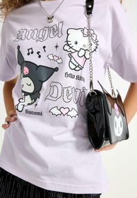 Fioletowa koszulka z grafikami Hello Kitty i Kuromi, ozdobiona błyszczącymi akcentami; w połączeniu z czarną torebką na ramię z lakierowanej skóry, mającą uszy nietoperza.