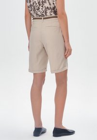 Beżowe szorty w luźnym fasonie z podwiniętymi nogawkami, wykonane z gładkiego materiału. Widoczne detale tylnej kieszeni; zestawione z czarnymi płaskimi butami.