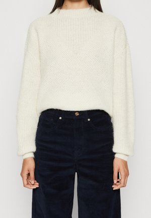 Frau trägt einen weißen, strukturierten Strickpullover mit langen Ärmeln, der in eine hoch taillierte schwarze Cordhose gesteckt ist.