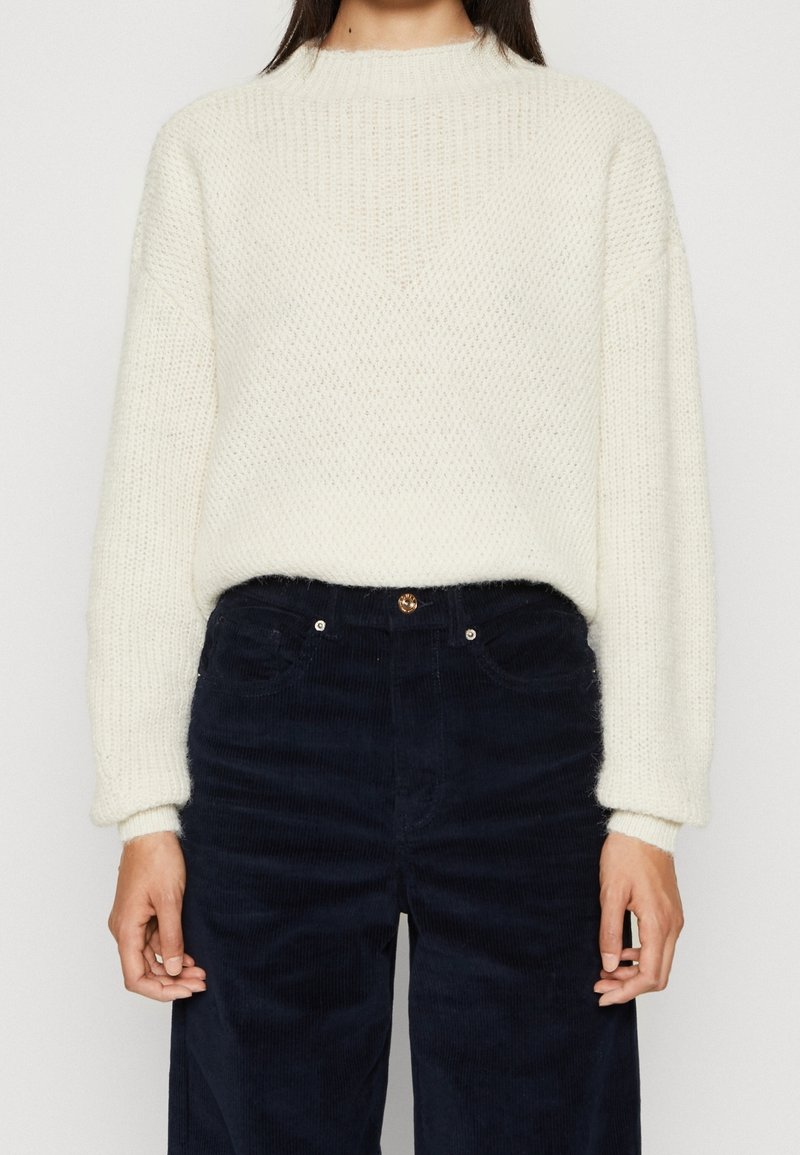 Femme portant un pull en maille texturée blanche à manches longues, rentrée dans un pantalon en velours côtelé noir taille haute.