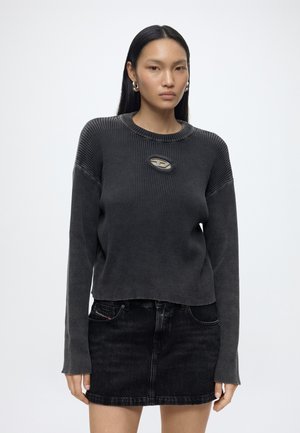 Femme aux cheveux noirs et raides portant un pull côtelé gris foncé avec une découpe ovale et un petit logo, associé à une jupe en jean noire.