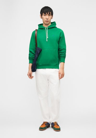 Moški stoji obrnjen naprej, oblečen v zeleno kapuco, bele hlače, večbarvne čevlje in ima torbo čez ramo s temno modrim pokrovom.