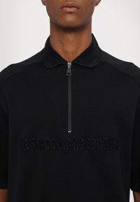 Svart polo-skjorte med kvart glidelås, strukturert krage og preget "VERSACE JEANSCOUTURE"-logo på forsiden. Glatt stofffinish.