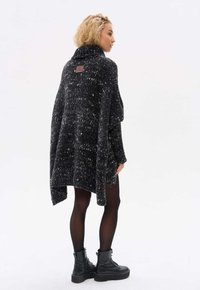 Przesadnie duży czarno-biały sweter w melanż, z wysokim kołnierzem i metką z tyłu, noszony na czarnych rajstopach i masywnych czarnych butach.