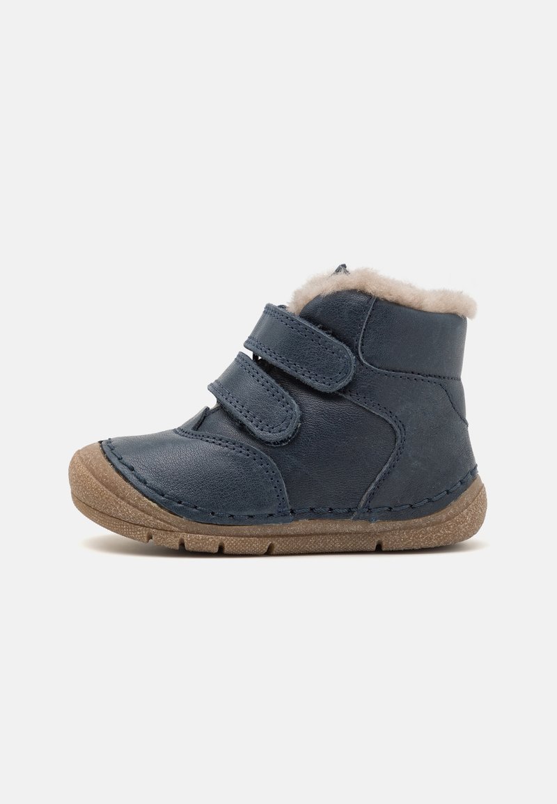 Botte en cuir bleu marine pour tout-petit avec deux sangles velcro, doublure beige duveteuse et semelle en caoutchouc marron texturée, orientée vers la gauche sur fond blanc.