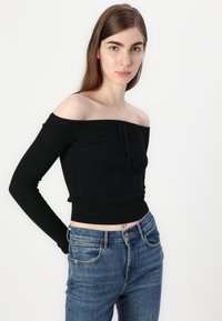 Pulôver preto de malha trançada, sem ombros, com mangas longas e um design cropped, combinado com jeans azuis de cintura alta.