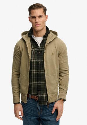 Junger Mann trägt einen hellbraunen Kapuzenpullover mit Reißverschluss über einem grünen karierten Hemd und einem weißen Unterhemd, kombiniert mit blauen Jeans und einem braunen Leder Gürtel.