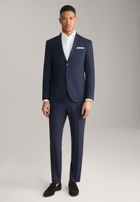 Abito blu navy con taglio slim fit, giacca a un bottone e due tasche. Camicia bianca fresca con colletto piegato. Mocassini neri.