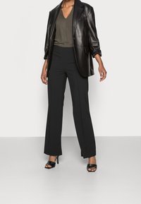 Woman wearing black wide-leg pants, black leather blazer, patterned V-neck top, and black open-toe high heel sandals, standing on gray floor.