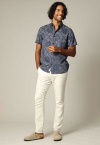 Korte mouwen, marineblauw overhemd met knopen en een bloemenpatroon, gecombineerd met witte broek en beige slippers van zacht materiaal.