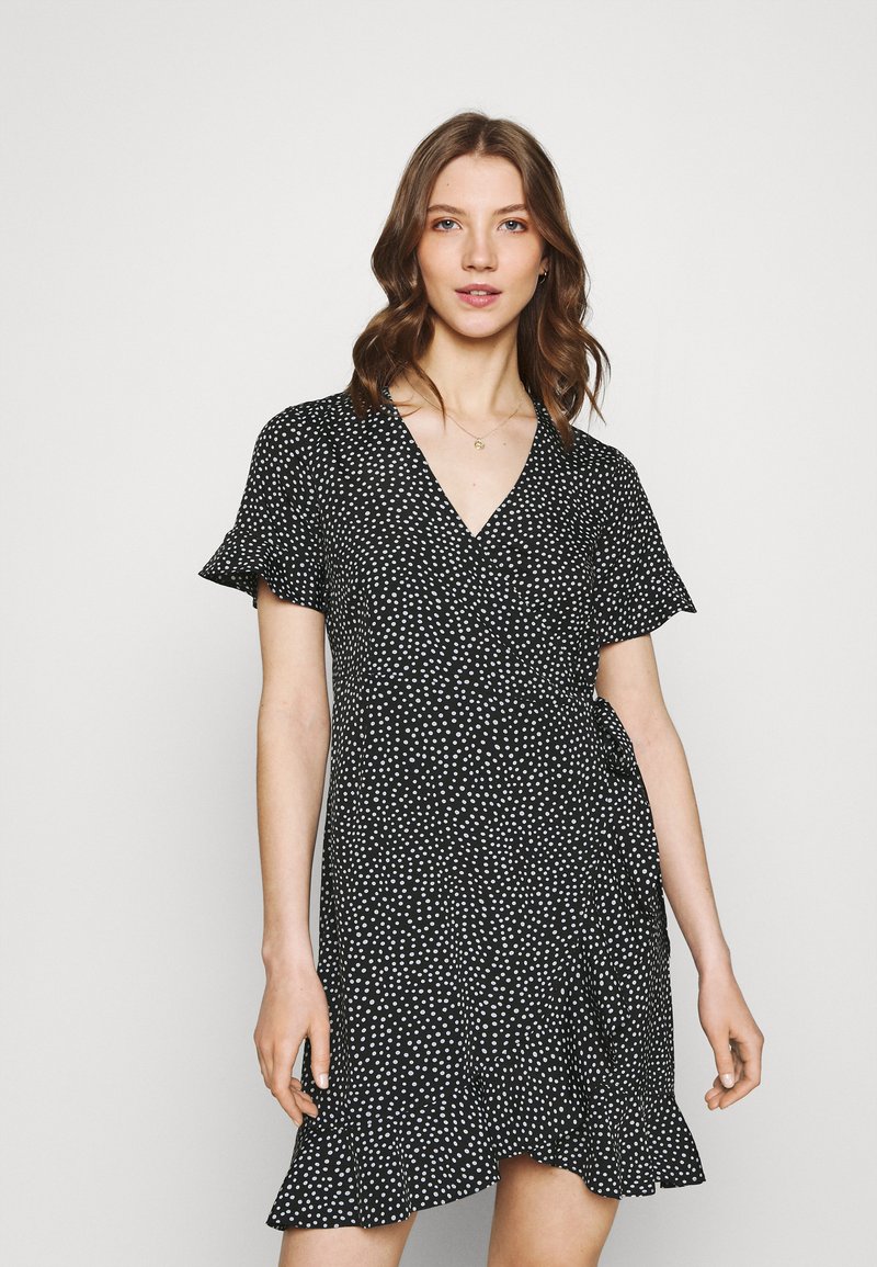 Vestido preto envolvente com bolinhas brancas, mangas curtas e esvoaçantes, decote em V, barra assimétrica e tecido leve. Adequado para ocasiões casuais.