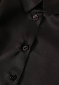 Černá saténová košile s hladkou texturou, vybavená špičatým límcem a třemi tmavými knoflíky s jemným logem značky.