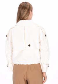 Weiße Jacke mit gerafften Ärmeln, großem Kragen und braunen Knopfdetails. Glatte Textur mit minimalen Designelementen.