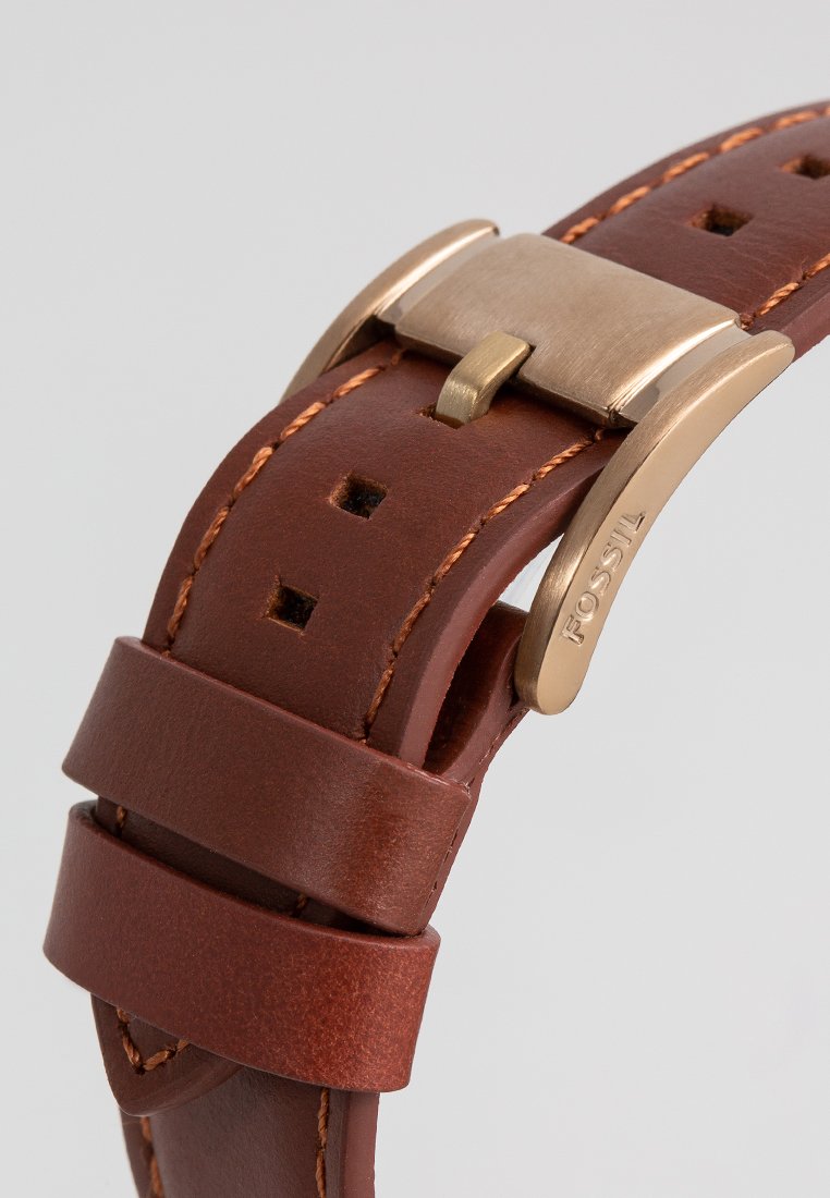 Correa de reloj de cuero marrón con detalles de costura, que cuenta con un cierre de metal y múltiples agujeros de ajuste. Superficie texturizada con un acabado pulido.