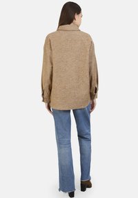 Bruine herringbone oversized blouse met een kraag, lange mouwen en knoopdetails, gecombineerd met lichtblauwe distressed jeans en bruine hakken.